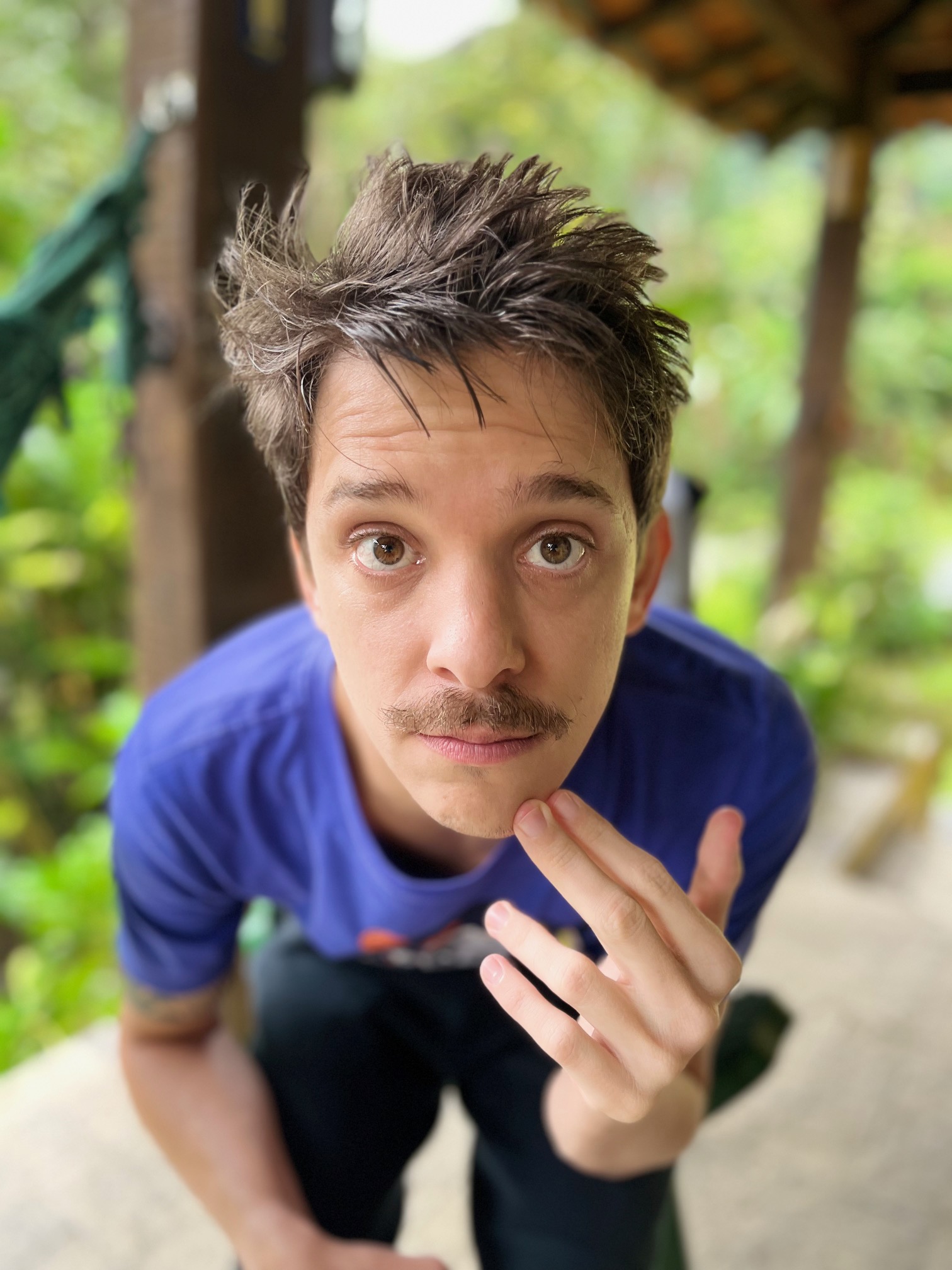 Homem com cabelo curto e bagunçado, bigode, usando uma camiseta azul, olhando diretamente para a câmera enquanto toca o queixo. Ao fundo, há uma área externa com vegetação.